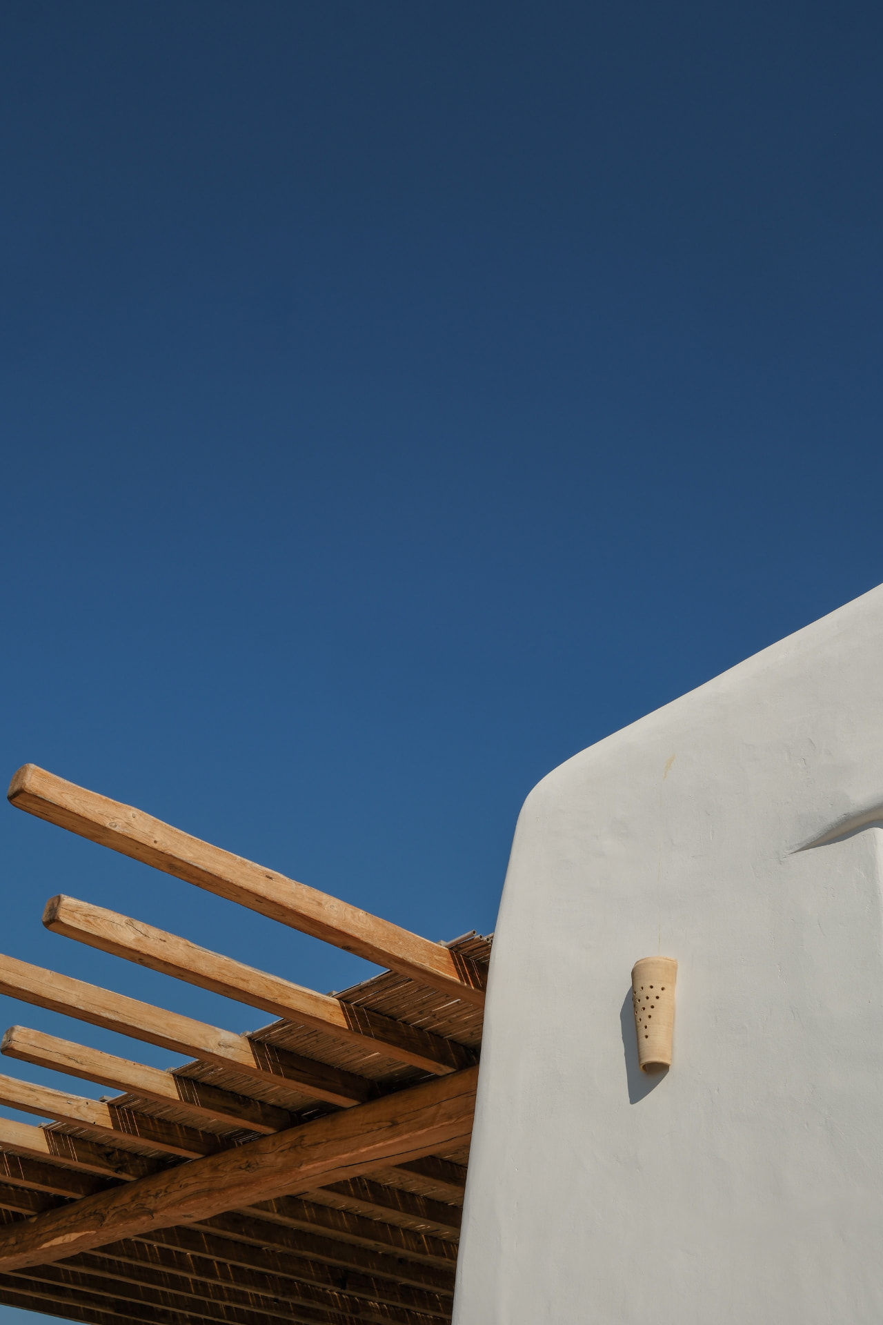 A smooth white wall and wall light beside a wooden pergola under a deep blue sky