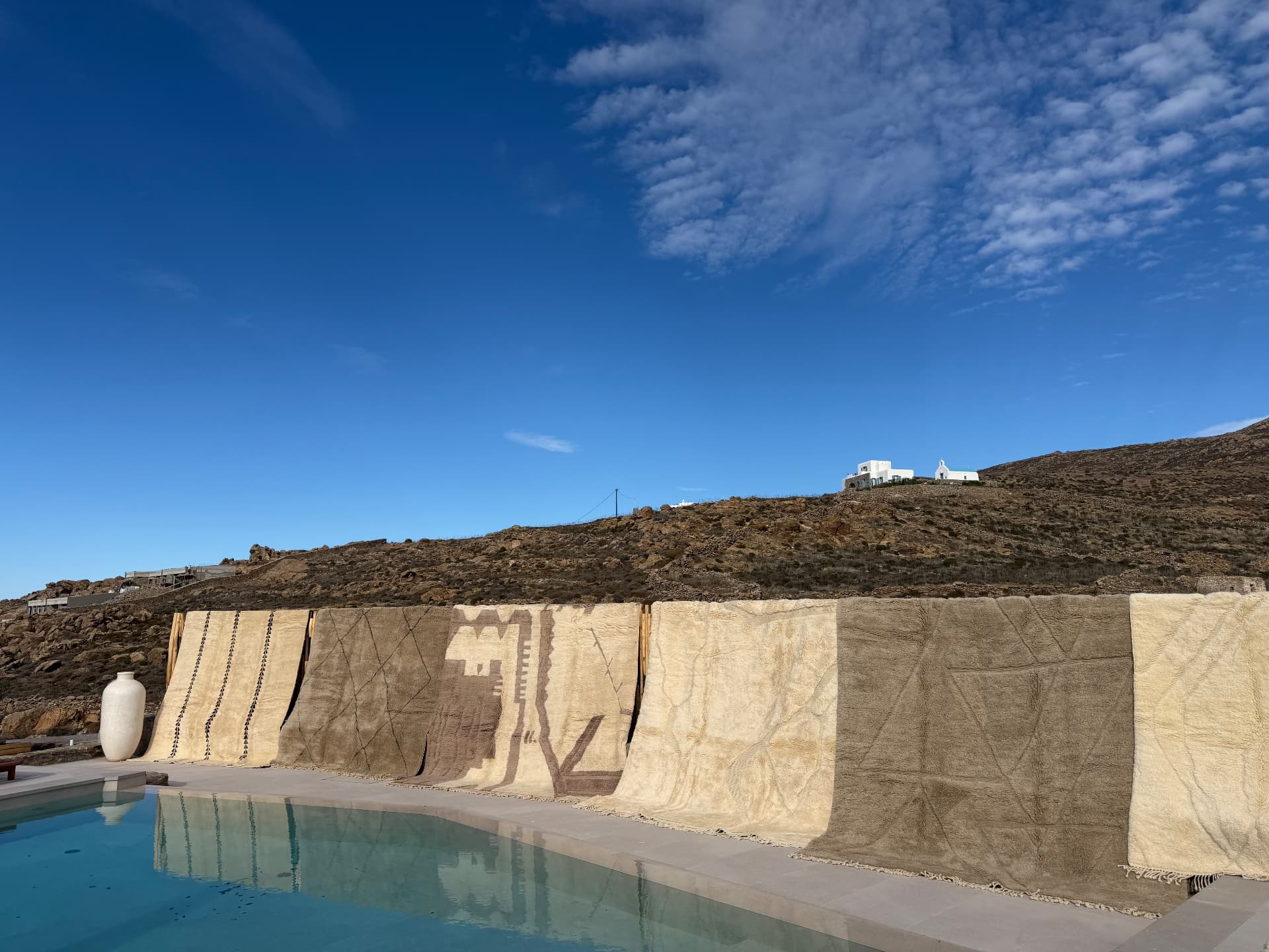 Handwoven rugs and tapestries draped over a low stone wall beside a turquoise pool, with a white ceramic urn nearby