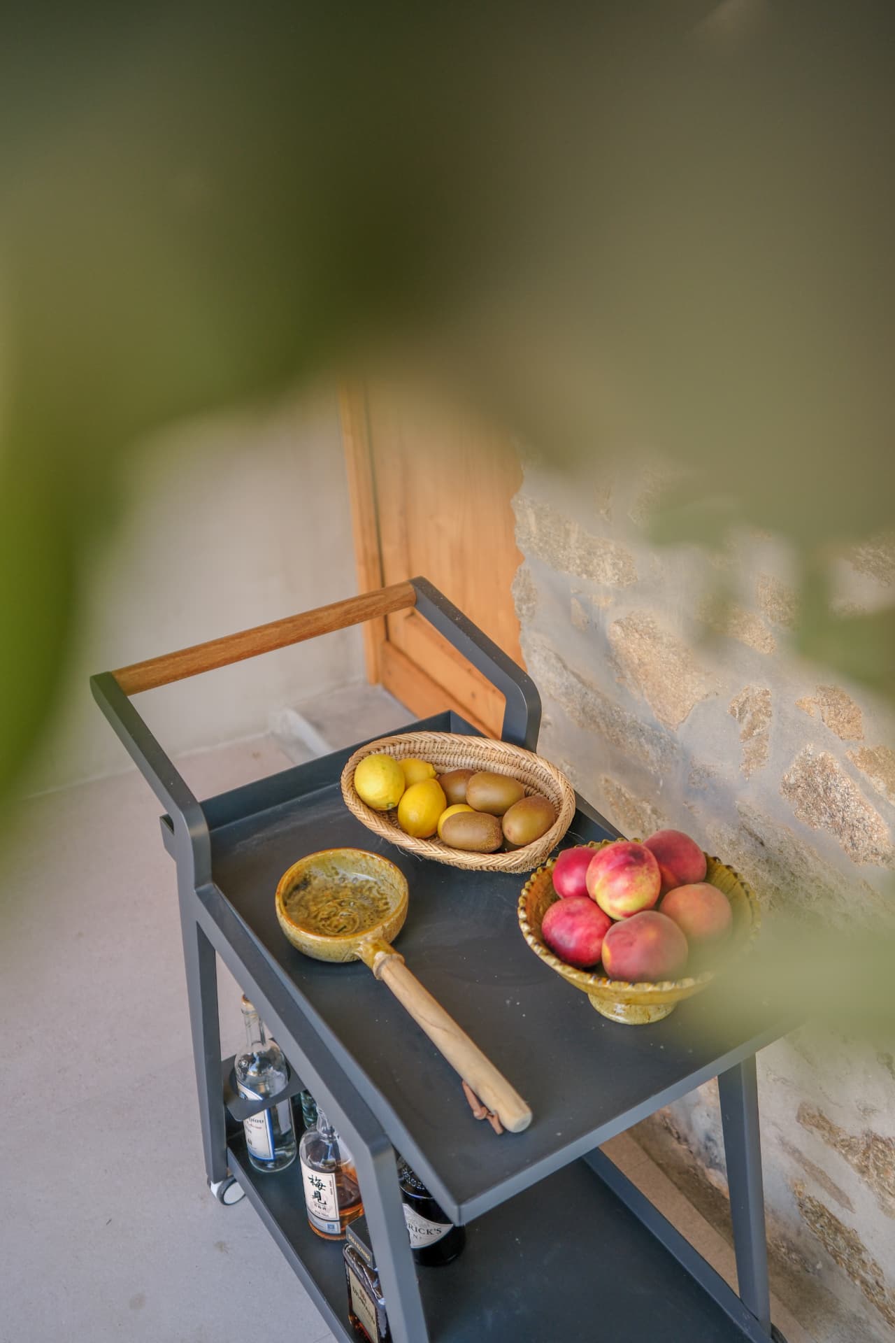 A dark grey two-tier bar cart against a stone wall, holding a basket of lemons and kiwis, a bowl of peaches and glass liquor bottles