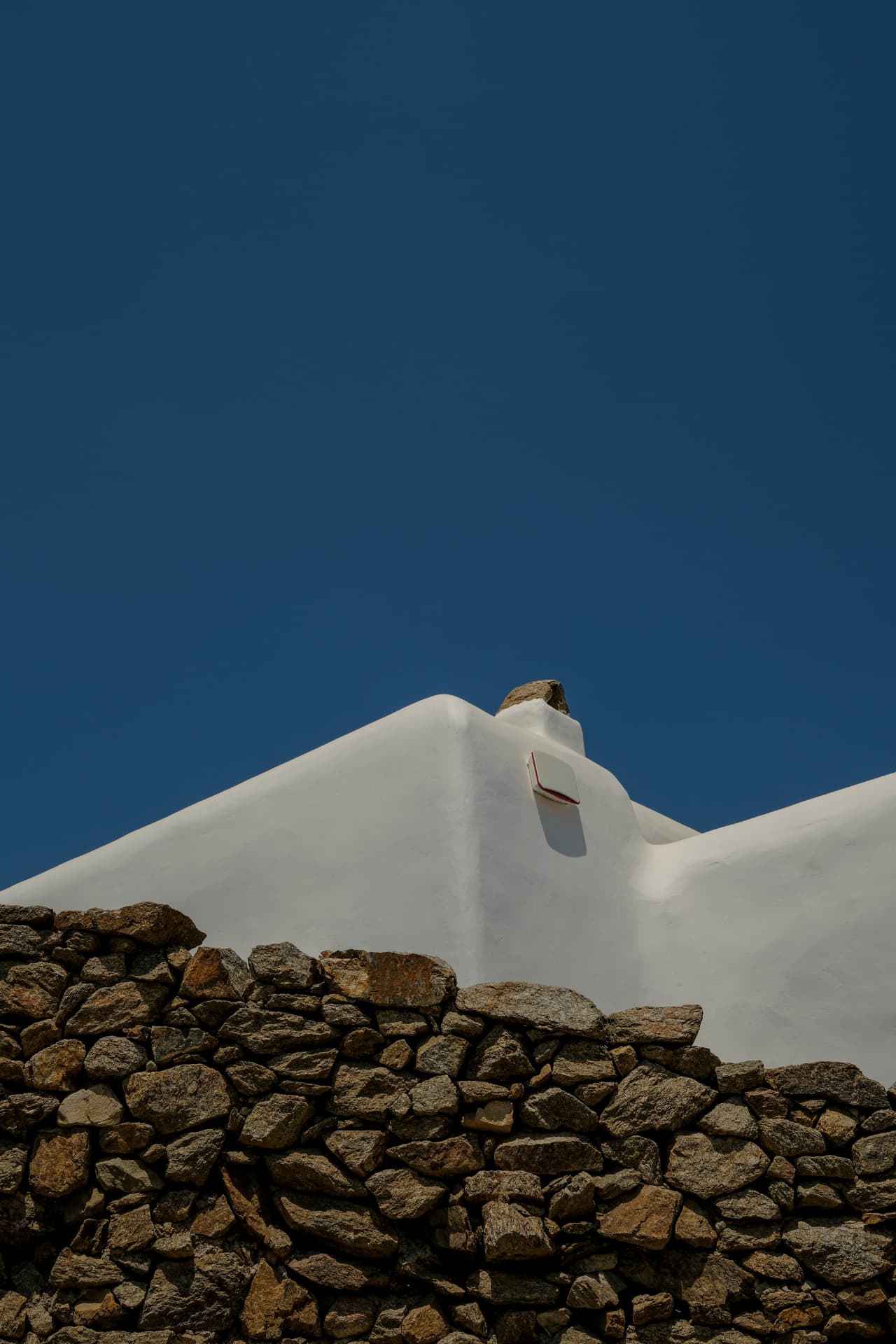 A white-washed building with rounded edges standing behind a rustic dry-stone wall under a deep blue sky
