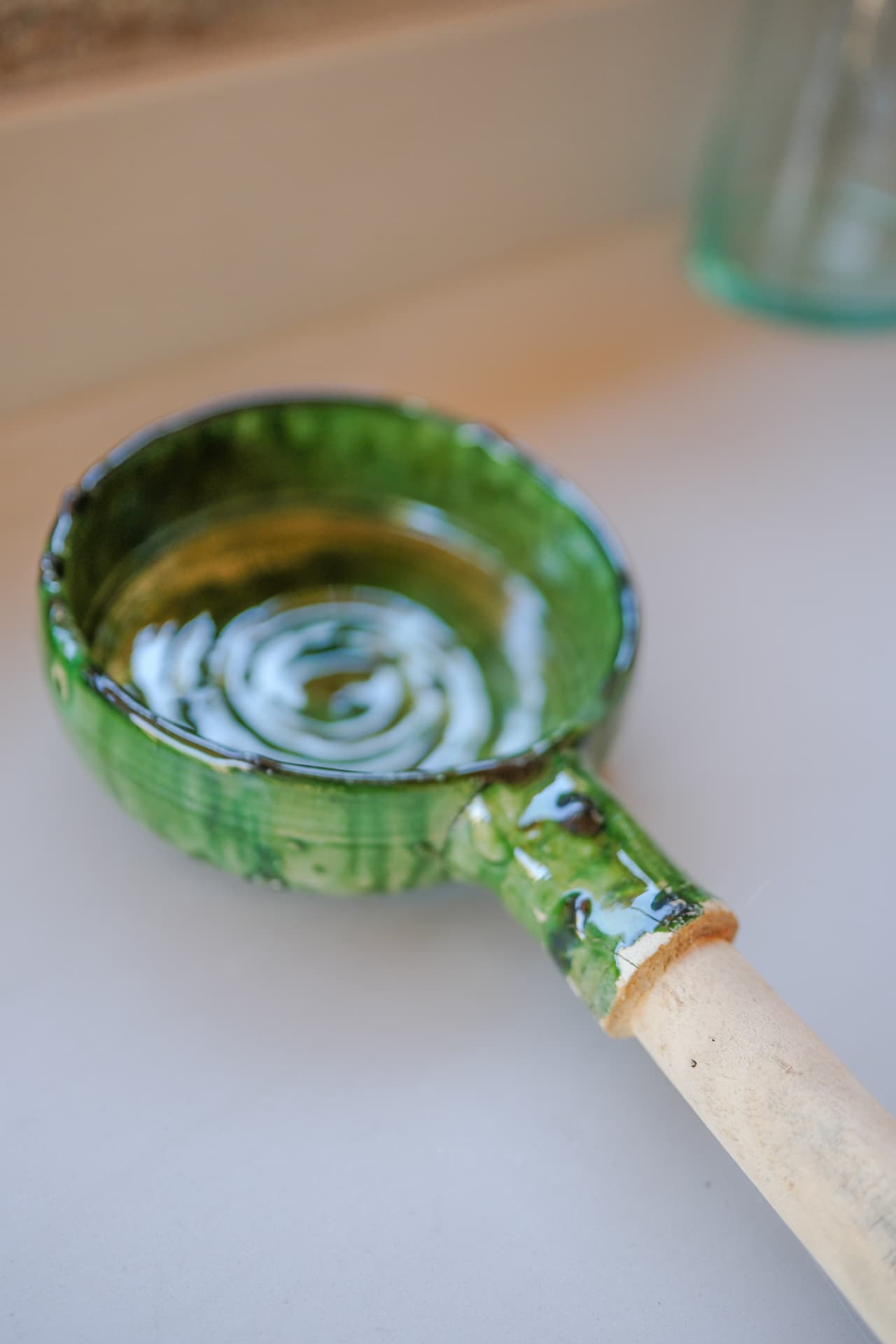 A green-glazed ceramic ladle with a wooden handle resting on a beige stone surface, water pooling in its bowl