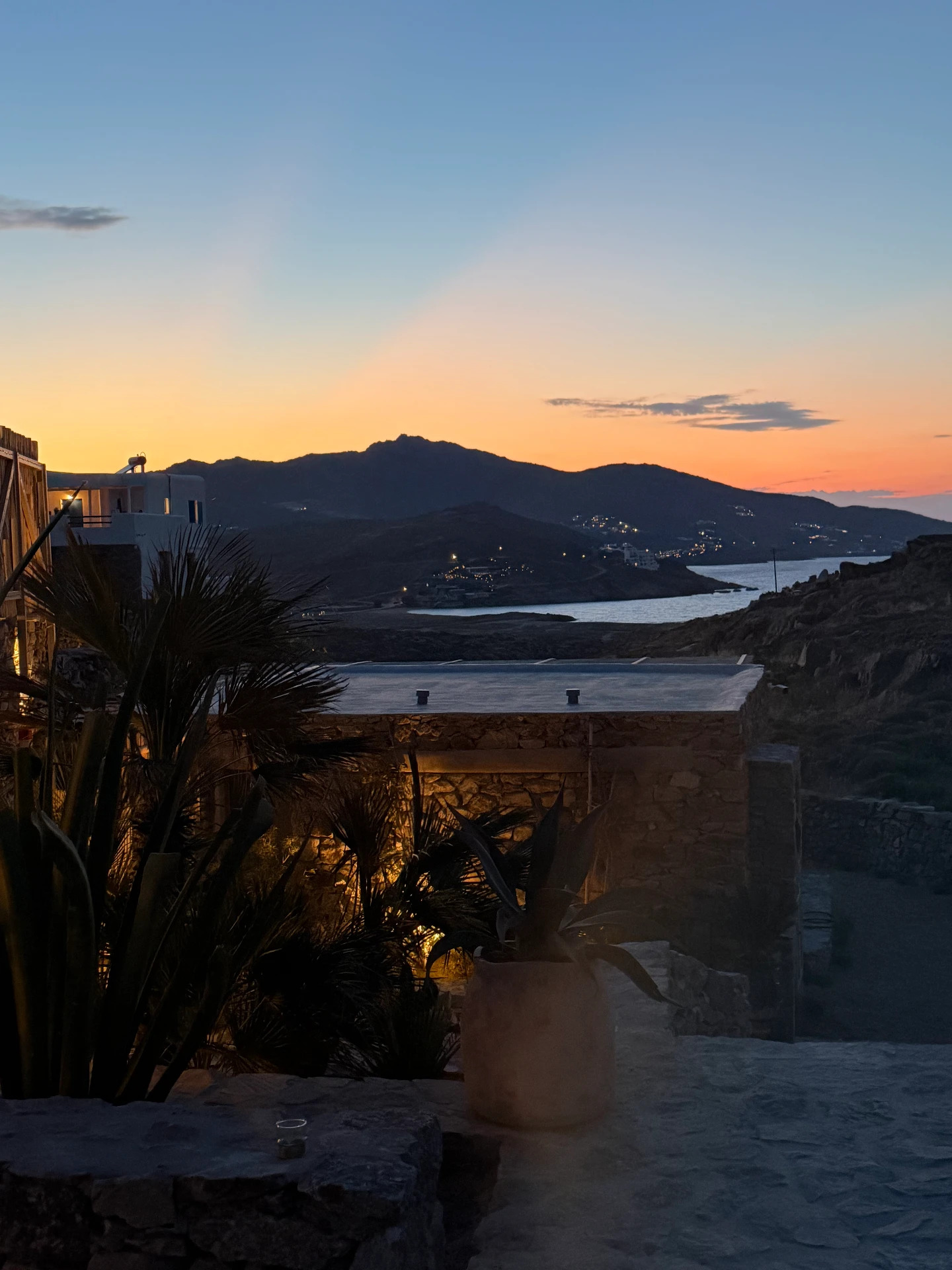 A serene sunset view from the stone terrace, silhouetted plants and a calm bay under an orange and blue sky