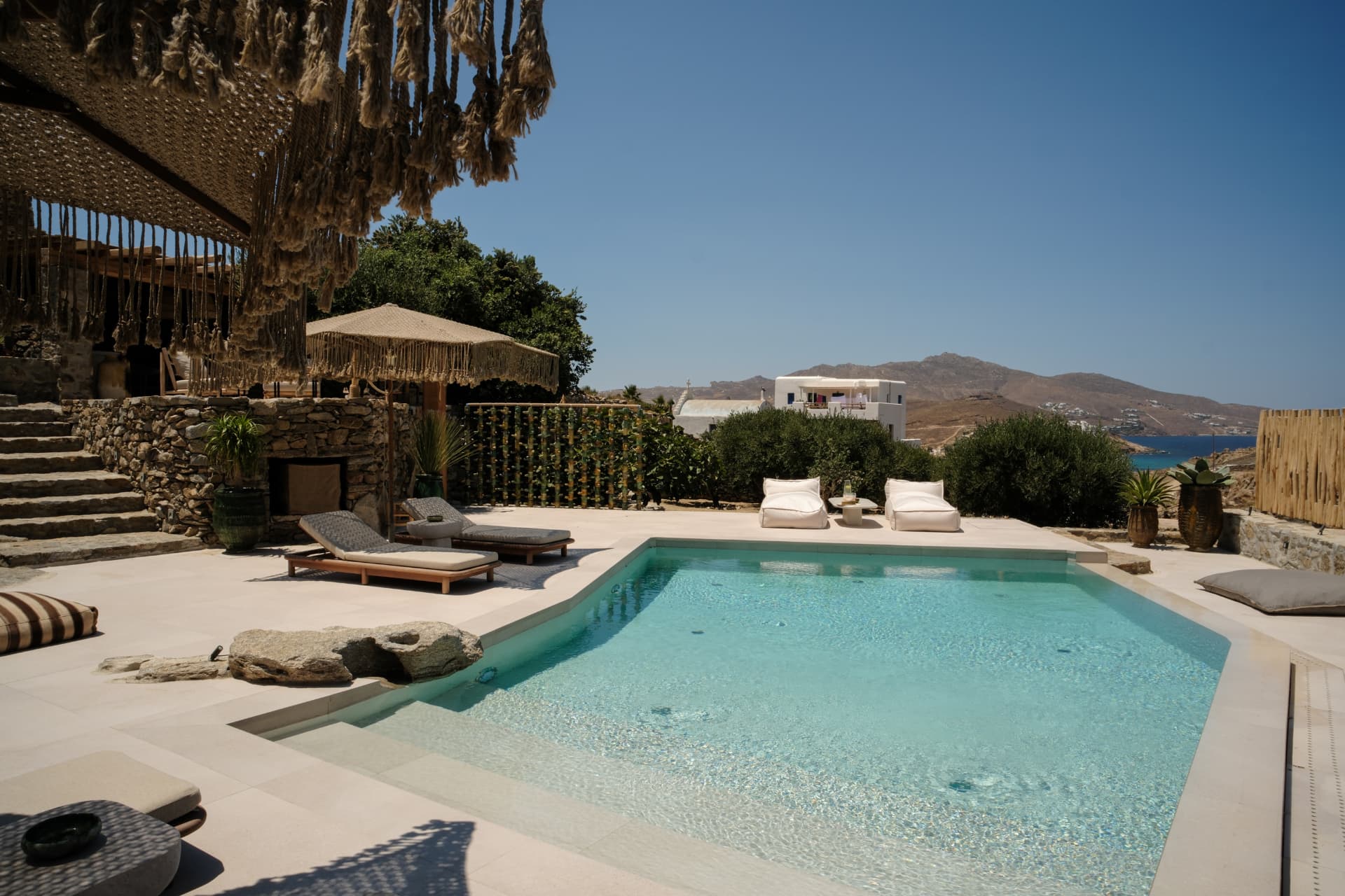 The pool at Villa Storia: turquoise water, wooden loungers under a macramé umbrella and the sea beyond