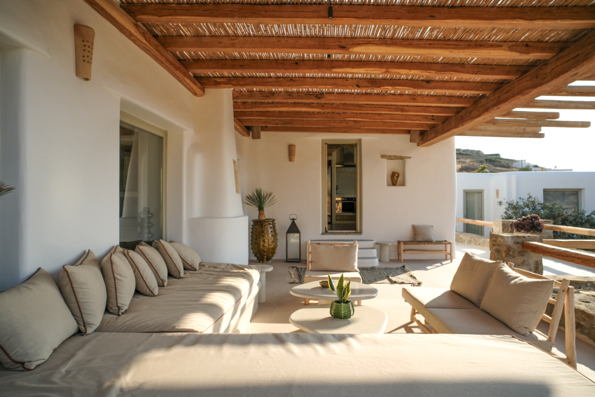 A shaded outdoor veranda with an L-shaped sofa, pale stone floor and warm wood pergola beams
