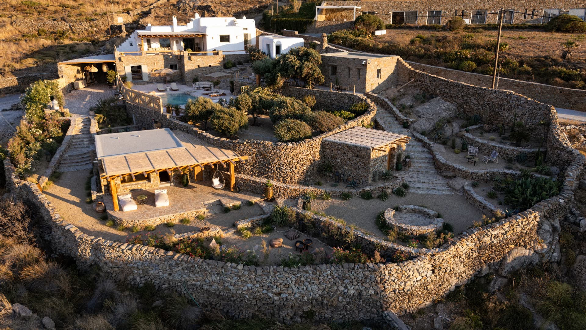 Aerial golden-hour view of Villa Storia's terraced gardens enclosed by curving dry-stone walls, with olive trees, cacti, a stone pergola house and the pool