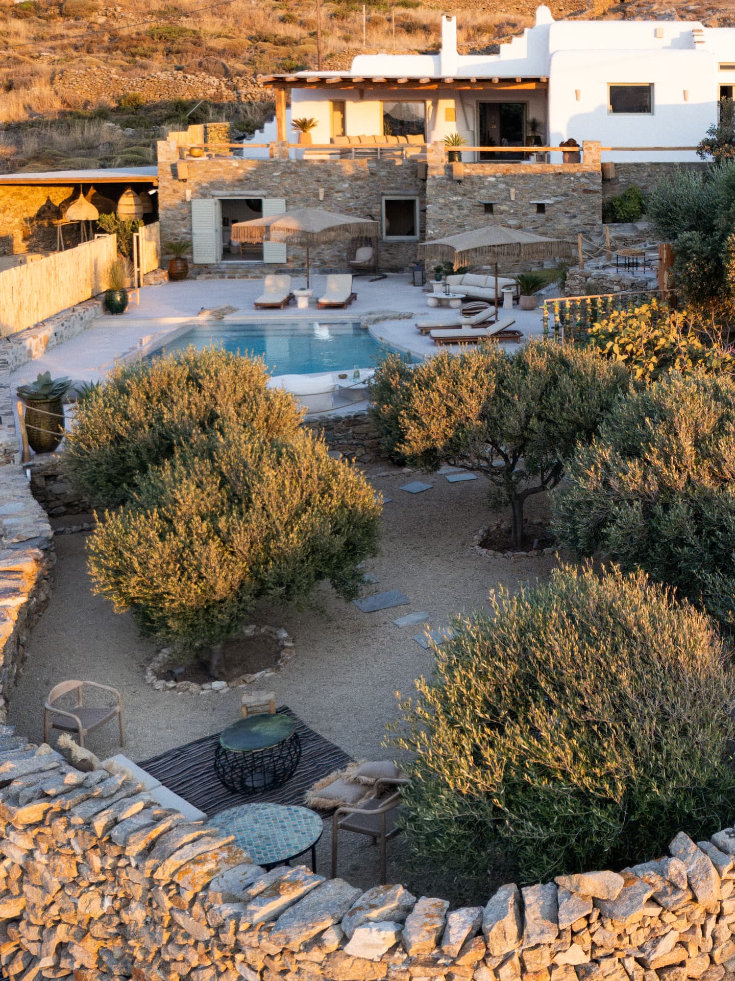 High-angle golden-hour view of Villa Storia: a curved dry-stone courtyard with olive trees and a seating area, a turquoise pool on a stone patio, and the white-washed villa terraces beyond