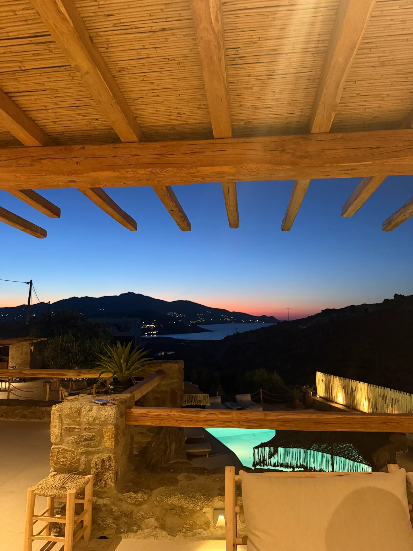 A peaceful dusk from the wooden-beamed patio, a glowing turquoise pool and distant coastal lights under a deep blue sky