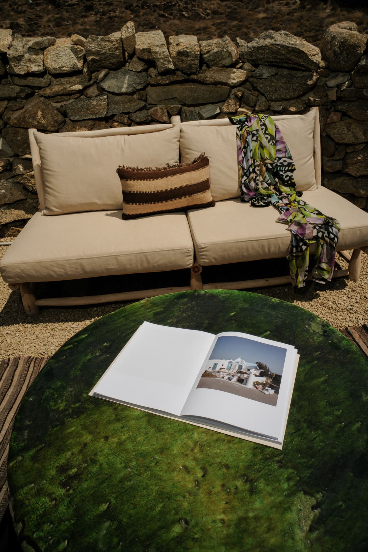 A cream sofa with a striped cushion and a patterned scarf against a dry-stone wall, an open book on a green glazed table