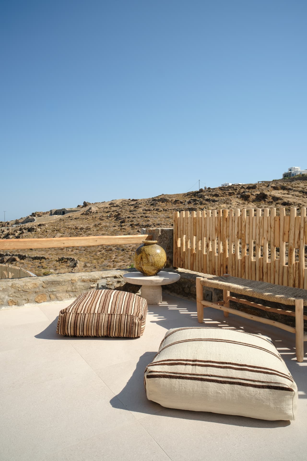 A minimalist roof terrace with striped floor cushions, a large ceramic vase and a vast rocky landscape beyond