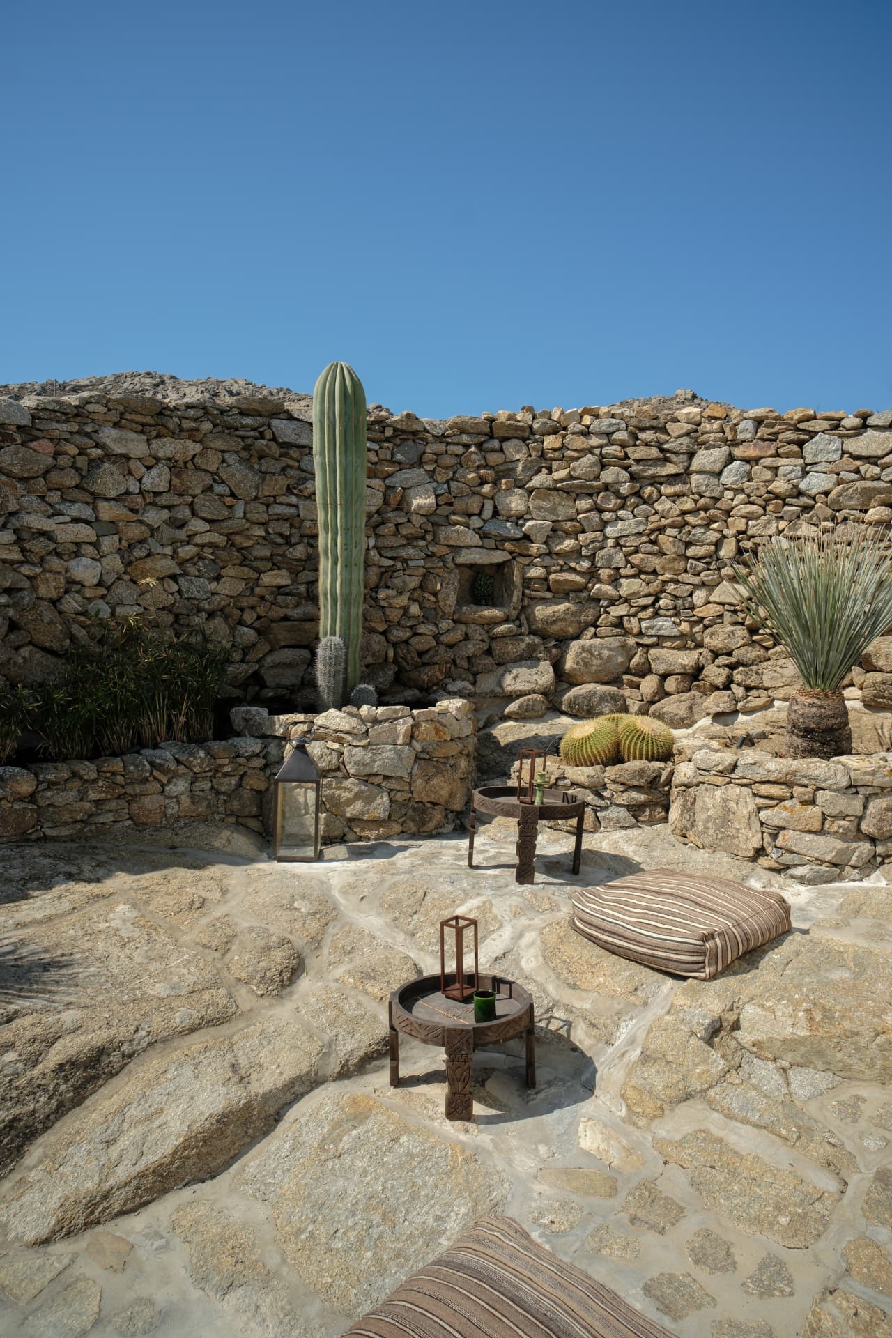 Patio corner with a tall green cactus, low wooden furniture and striped cushions within a stone-walled enclosure under a blue sky