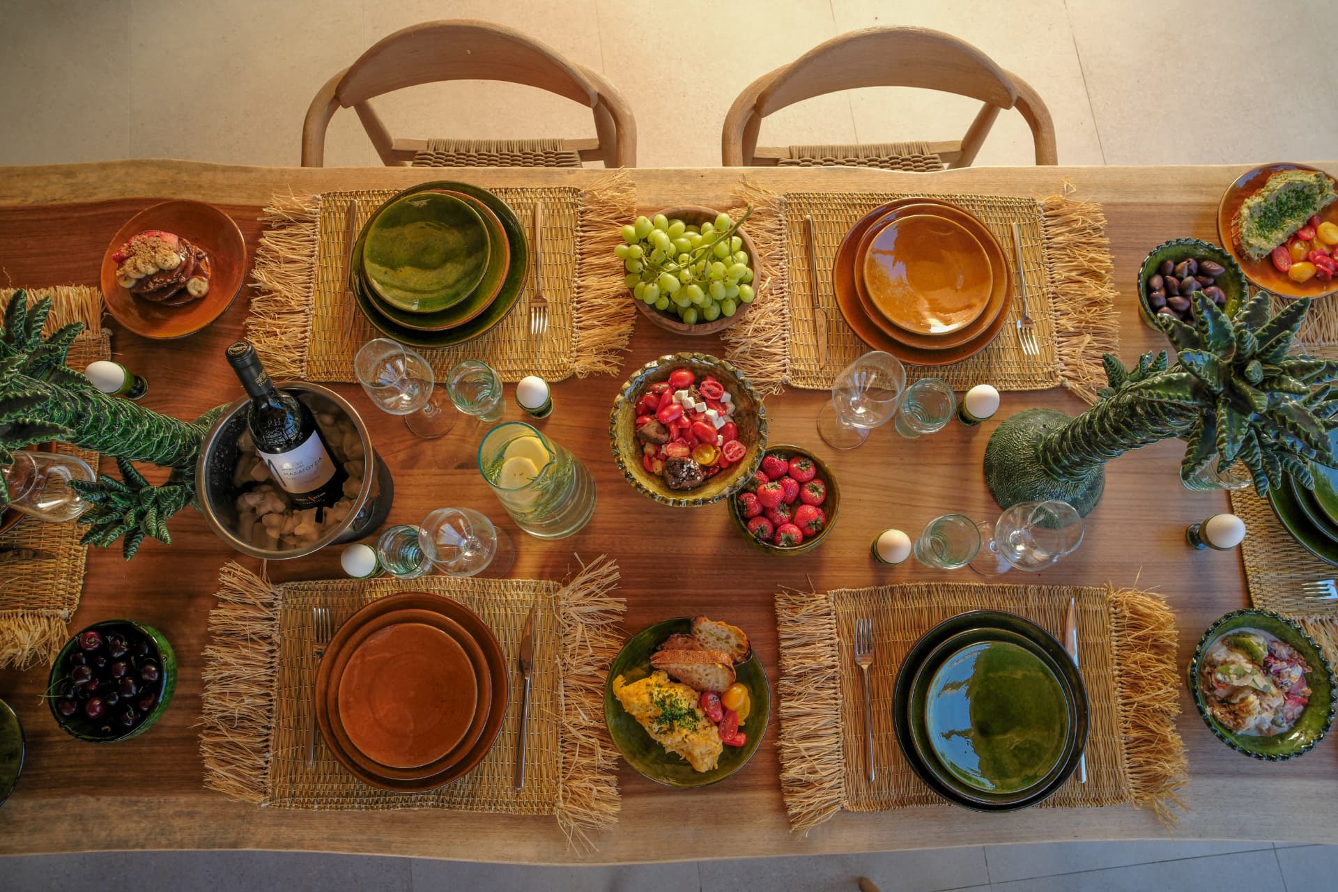 Top-down view of the wooden outdoor table fully set, straw placemats, green and burnt-orange ceramics, bowls of fruit and a wine bucket