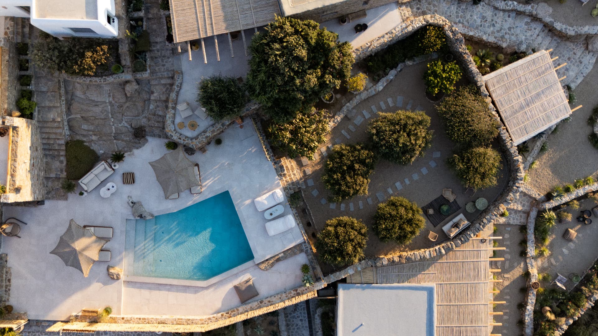 Straight-down aerial view of the pool patio and an enclosed olive garden divided by a curved dry-stone wall, with stepping stones across gravel