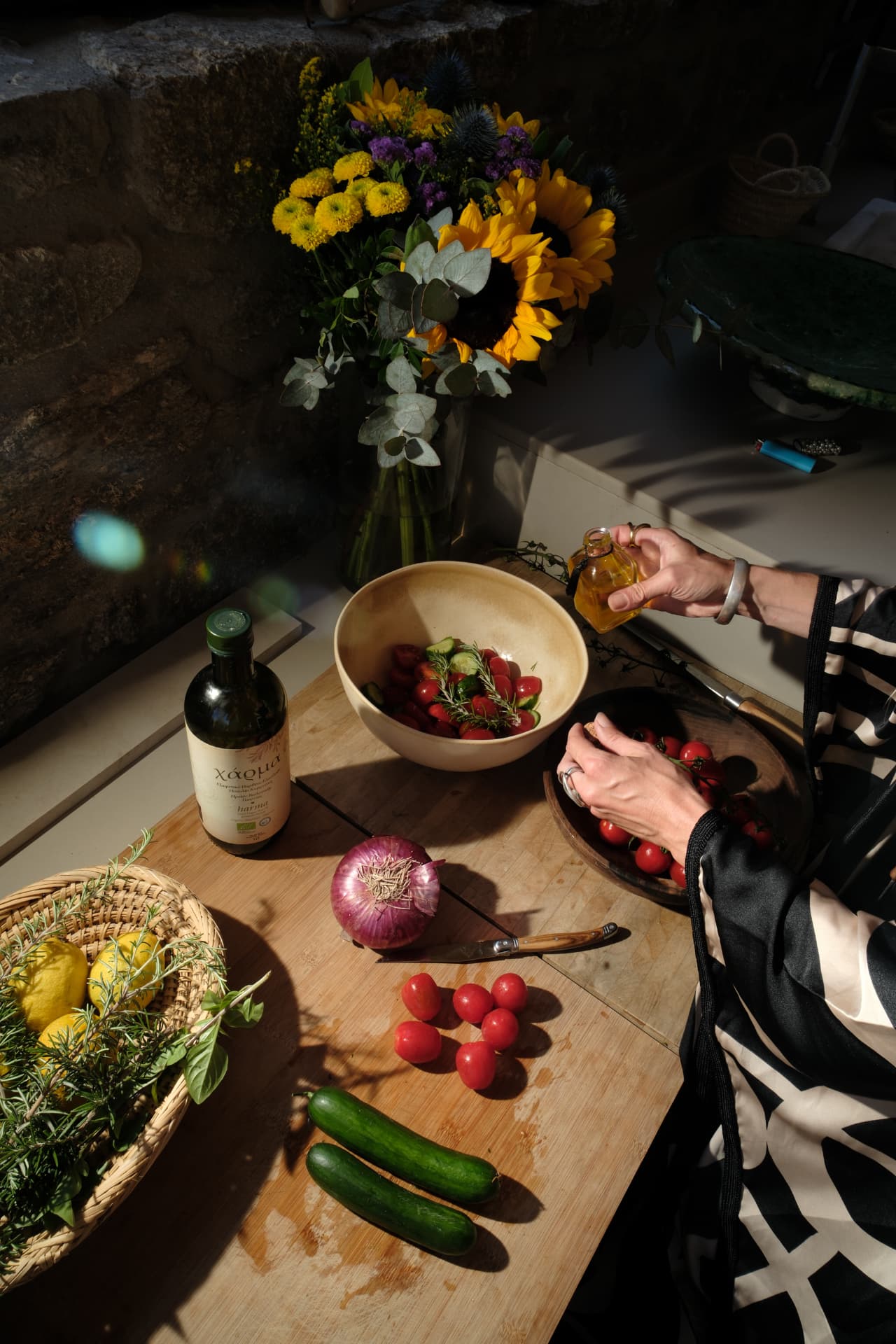 Hands pouring olive oil into a ceramic salad bowl on a wooden counter against a stone wall, with lemons, cucumbers and a bouquet of sunflowers