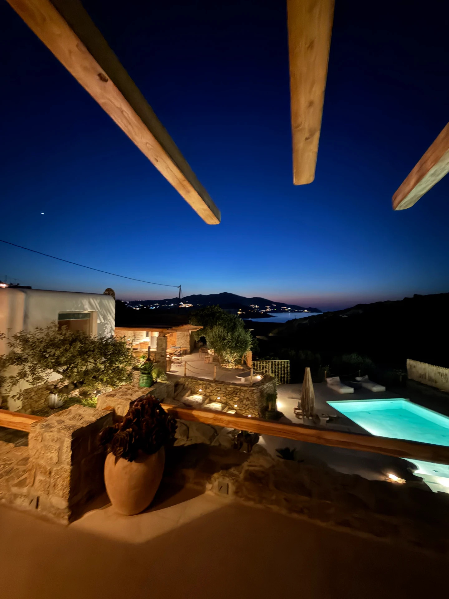 The illuminated pool and stone terraces seen beneath wooden pergola beams against a deep night sky and distant coastal lights