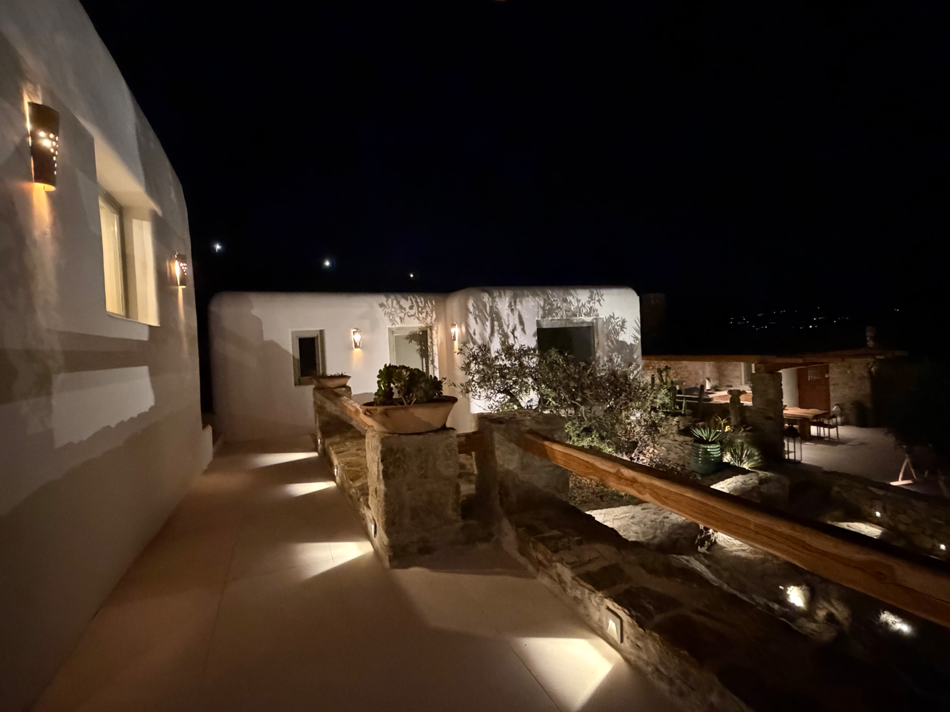A warmly lit outdoor walkway and patio between white Mediterranean buildings and stone walls under a dark sky
