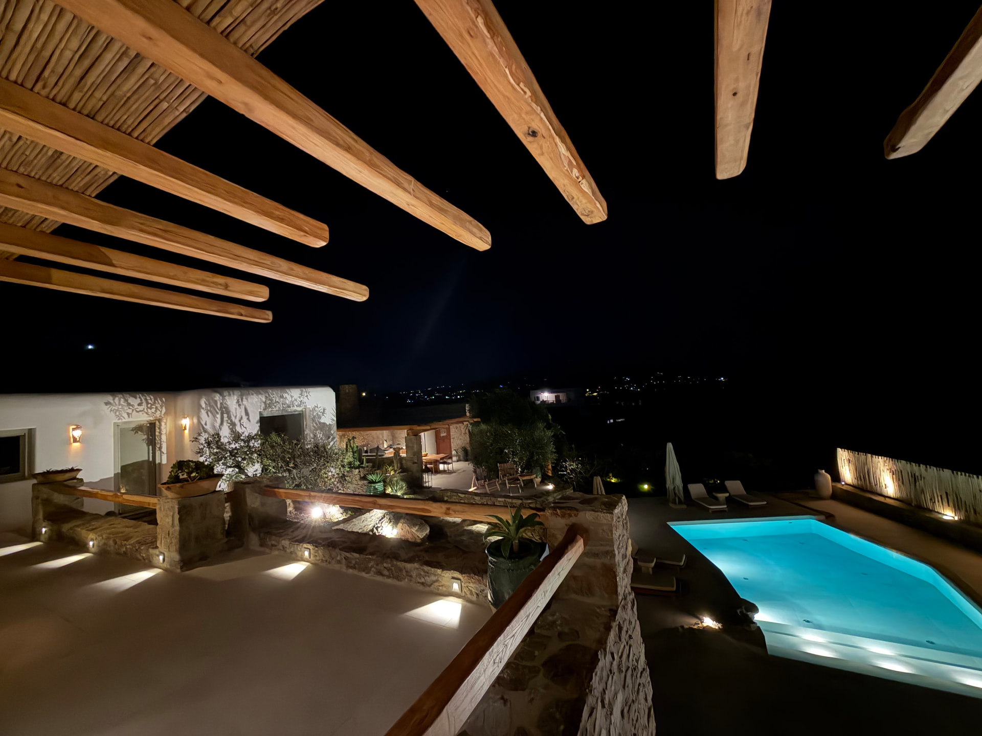 Night-time view through wooden pergola beams over the illuminated swimming pool, with distant town lights beyond