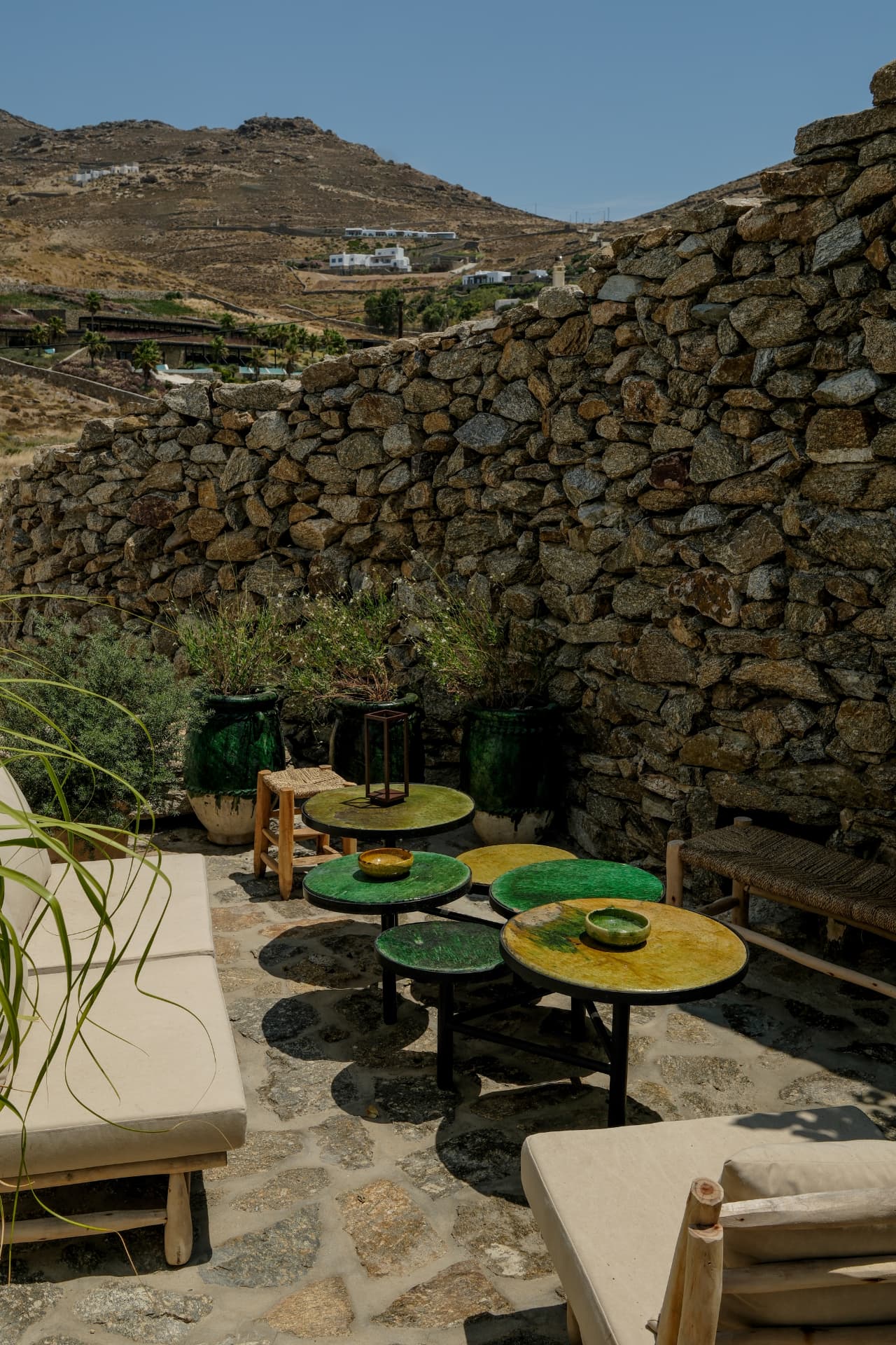 Patio seating with green and yellow glazed tables, woven stools and cream-cushioned seating against a thick stone wall