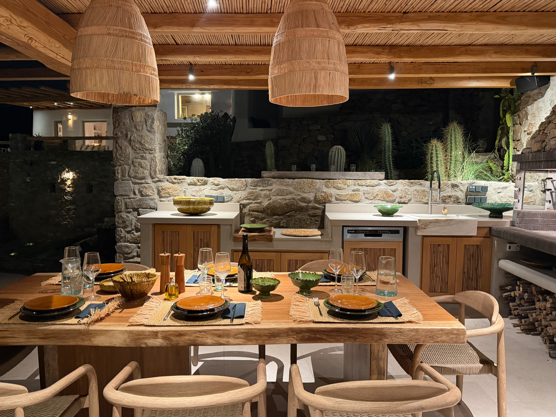 The outdoor dining table set beneath two large wicker pendant lights, beside the stone-walled kitchen at night