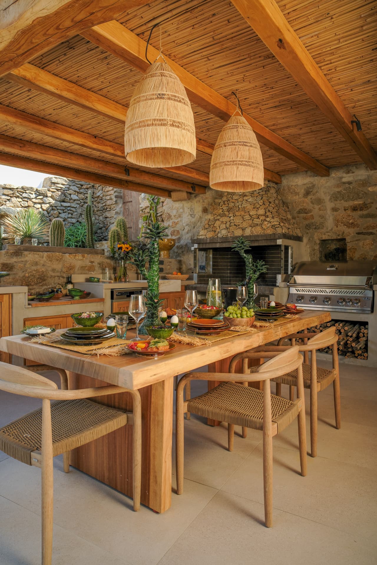 The outdoor kitchen with a large light-wood table, woven bell pendant lights, stone walls, a built-in grill and a wood-filled stone fireplace