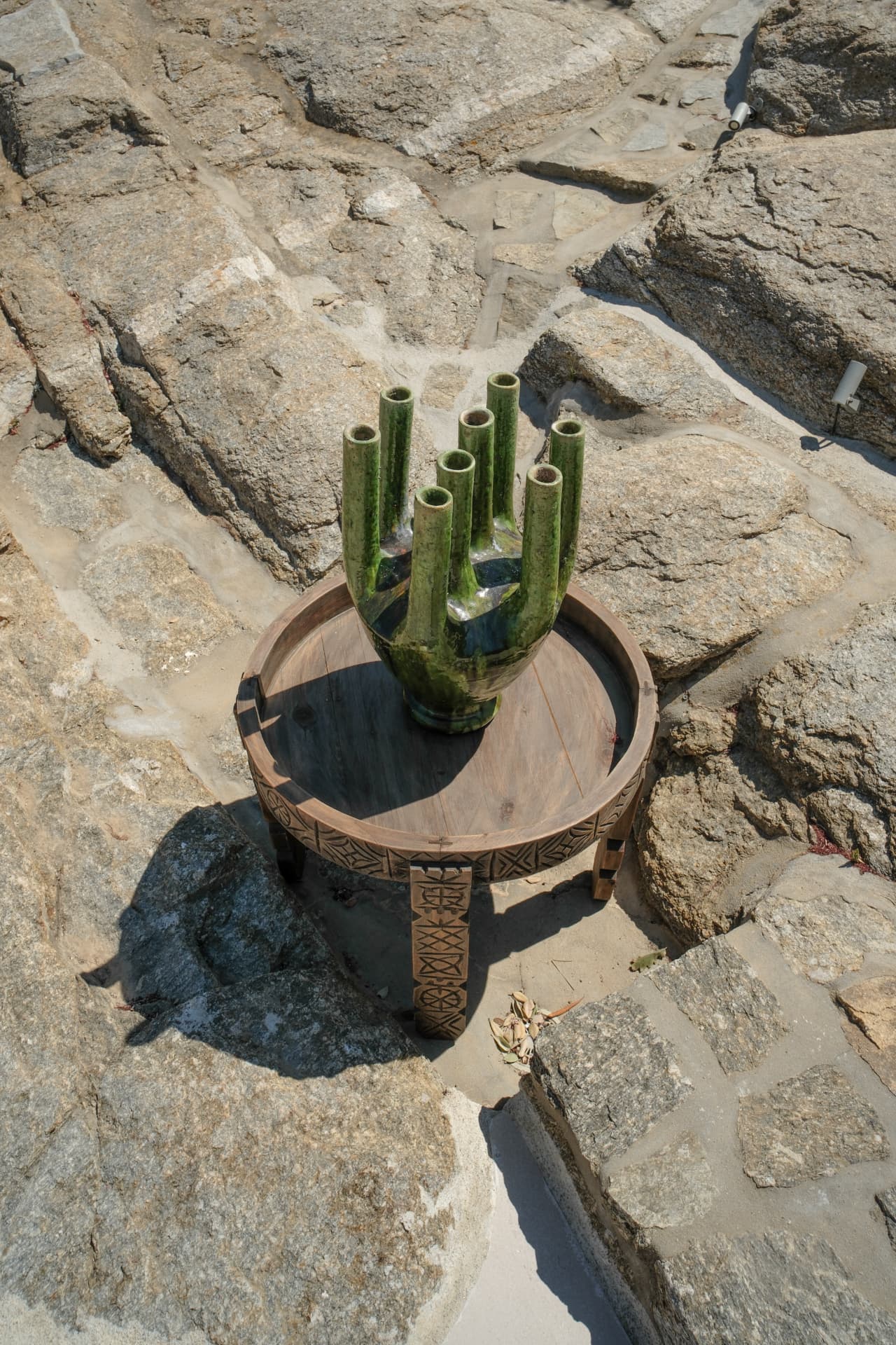 A green glazed multi-spouted ceramic vase on a carved wooden tray table set among granite rocks
