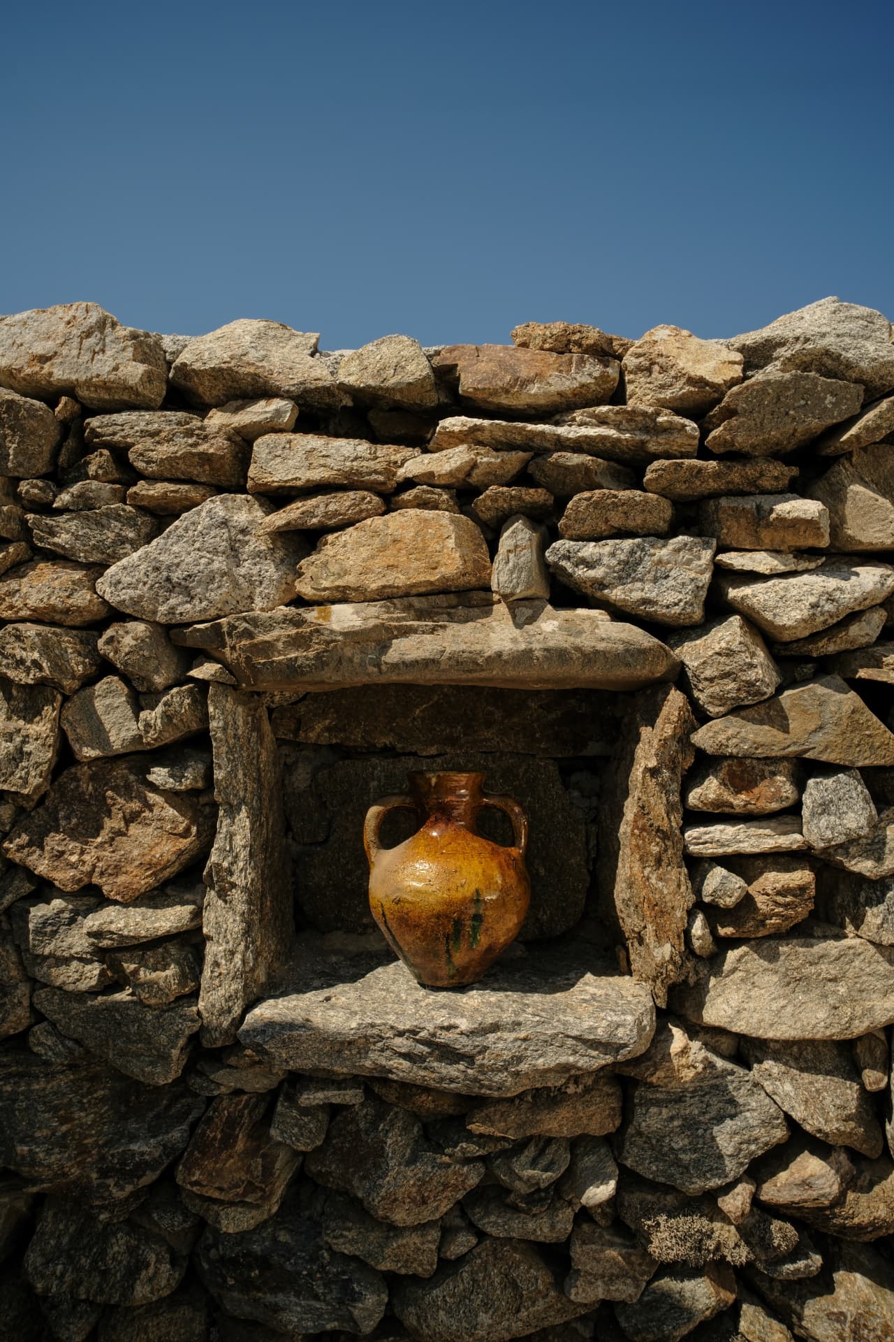 A glossy amber earthenware jar inside a rectangular stone niche built into a light-brown rock wall
