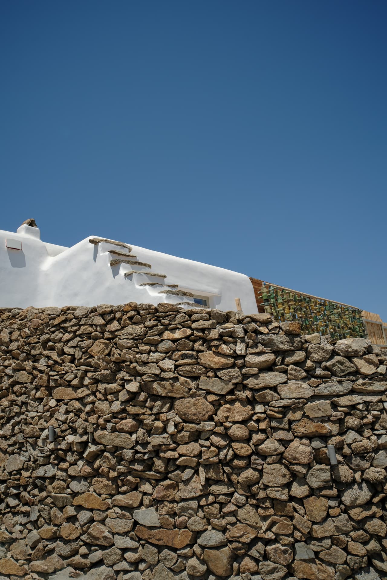 A whitewashed Cycladic building with stone steps built into its side and a decorative wall of green glass bottles, behind a textured dry-stone wall under a deep blue sky