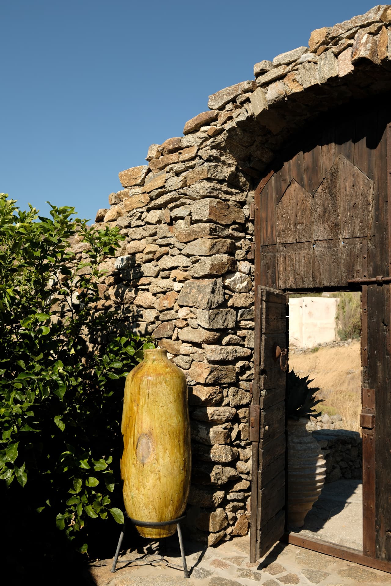 A tall yellow-glazed ceramic urn beside a partly open arched wood door set in a rustic stone wall