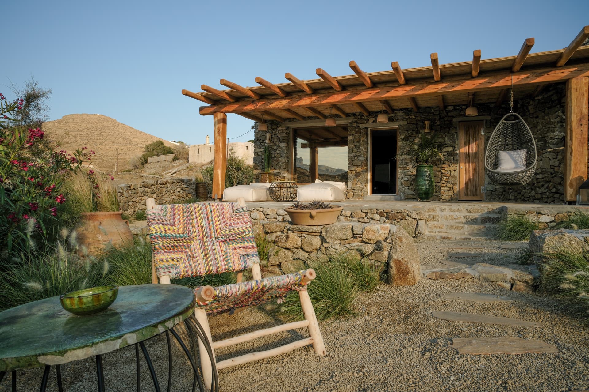 A woven multicoloured chair and green glazed table on gravel before a stone house with a wooden pergola and hanging egg chair