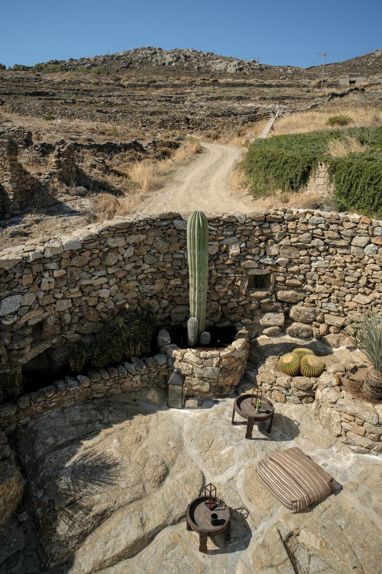 High-angle view of a circular stone-walled terrace with a tall cactus, low tray tables and a winding dirt road